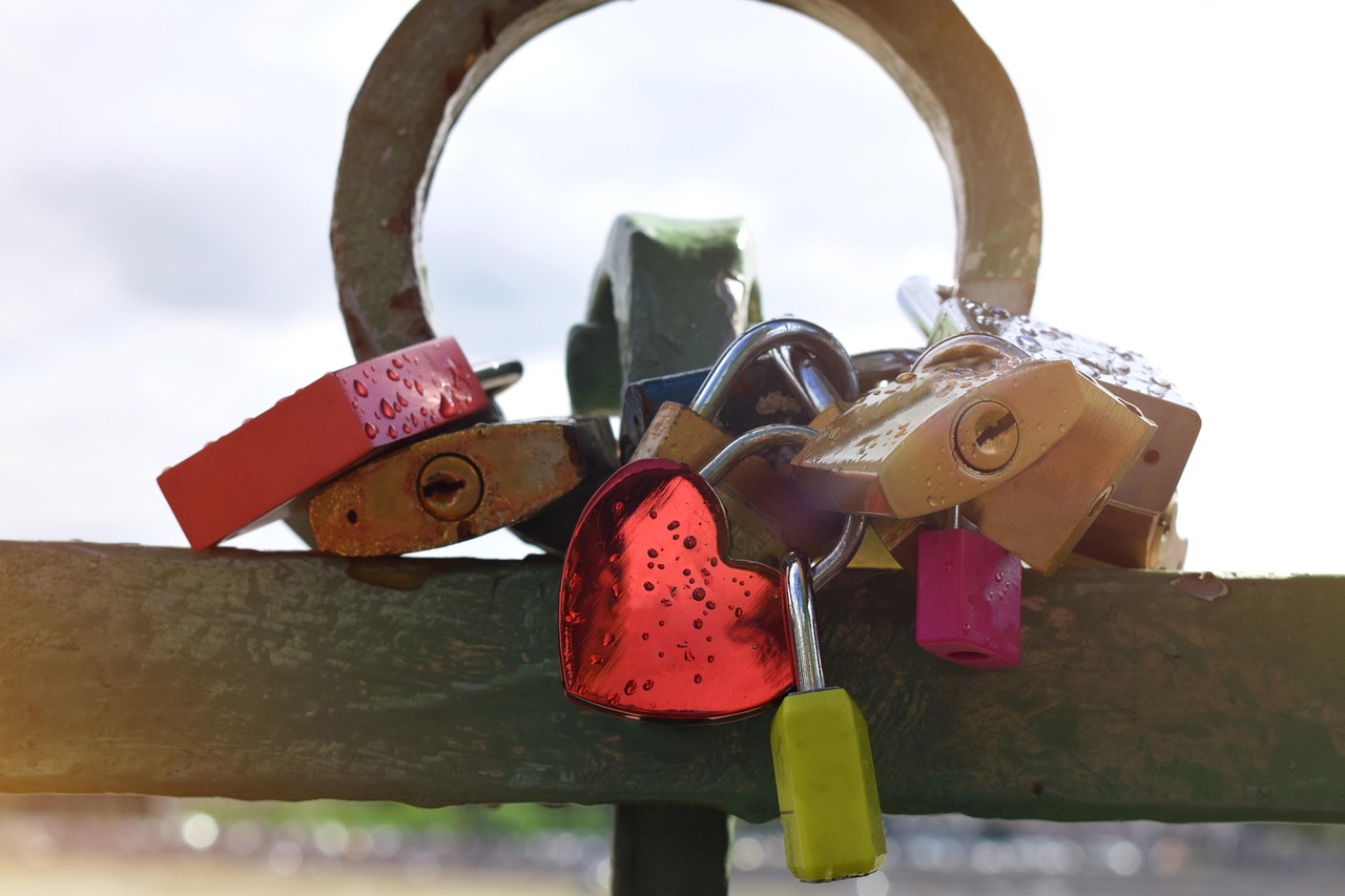 Mehrere Schlösser in verschiedenen Farben, die an einem Geländer festgemacht sind. Eins der Schlösser hat eine rote Herzform mit paar Tropfen darauf.